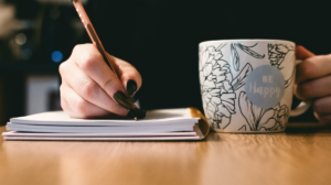 Une personne écrit dans un cahier tout en tenant une tasse en céramique « Be Happy » — une image qui évoque l’inspiration, la créativité et l’esprit des jams de création présentés dans cet article.